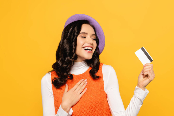 Smiling brunette woman in beret holding hand on chest and looking at credit card isolated on yellow - Photo, Image