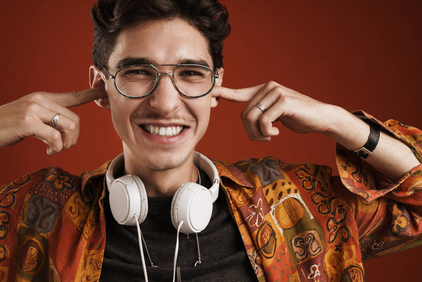 Close up of a smiling young teenage boy wearing glasses standing isolated over red background, wearing headphones, laughing - Foto, afbeelding