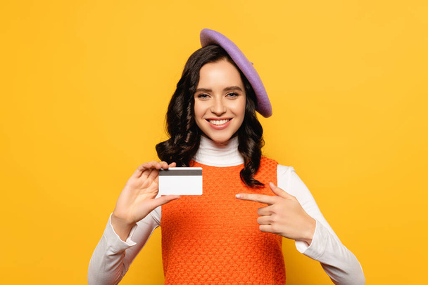 Smiling brunette woman in beret looking at camera while pointing with finger at credit card isolated on yellow - Zdjęcie, obraz