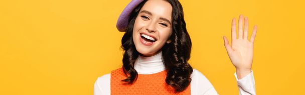 Happy brunette woman in beret with waving hand looking at camera isolated on yellow, banner - Foto, Imagen