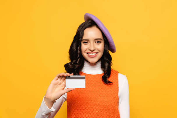 Smiling brunette woman in beret looking at camera while showing credit card isolated on yellow - Foto, Imagen