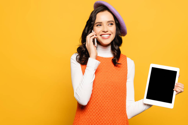 Happy brunette woman in beret talking on smartphone while holding digital tablet with blank screen isolated on yellow - Foto, immagini