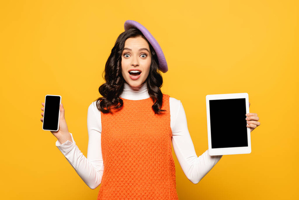 興奮ブルネット女性でオープン口身に着けているベレー帽とショースマートフォンとデジタルタブレット孤立した上黄色 - 写真・画像