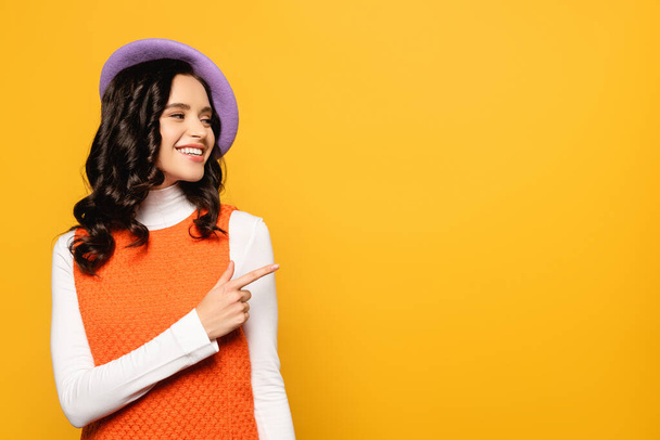 Happy brunette woman in beret looking away while pointing with finger isolated on yellow - Фото, зображення