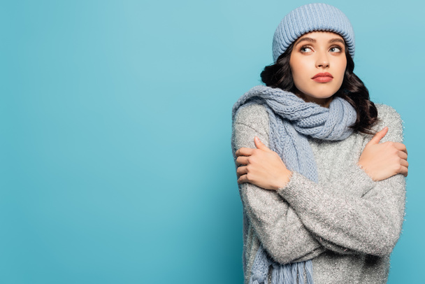 Brunette woman in winter outfit hugging herself while looking away isolated on blue - Photo, Image