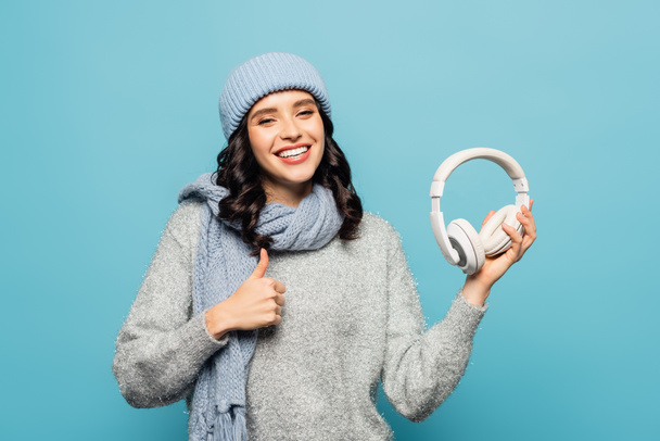 Happy brunette woman in winter outfit holding headphones and showing thumb up isolated on blue - Foto, afbeelding