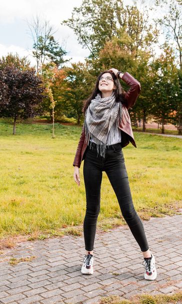 junges Mädchen in brauner Jacke und schwarzer Jeans steht an einem sonnigen Herbsttag auf der Gasse im Stadtpark - Foto, Bild