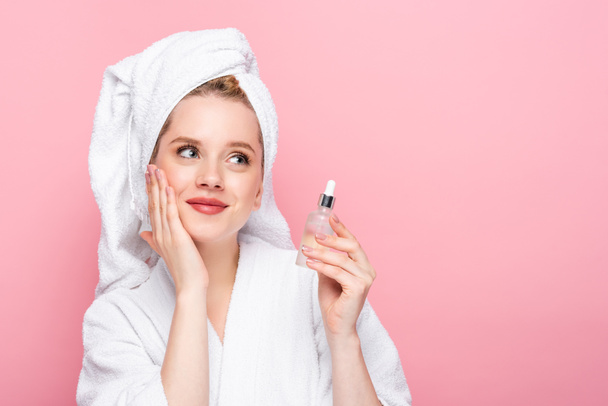 dreamy young woman in bathrobe with towel on head holding serum isolated on pink - Фото, зображення