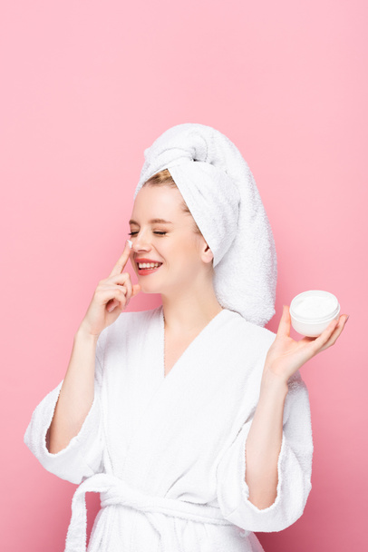 heureuse jeune femme en peignoir serviette sur la tête tenant la crème cosmétique et l'appliquant sur le nez isolé sur rose - Photo, image
