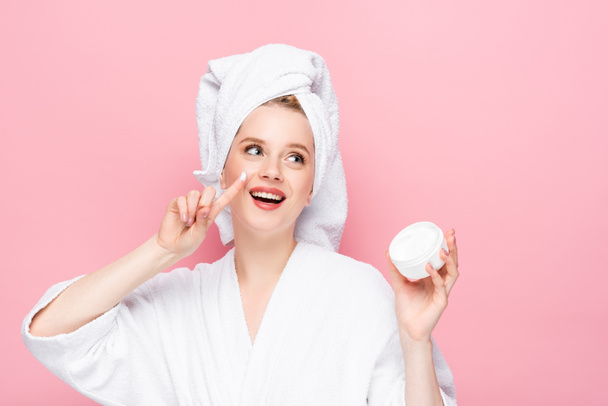 heureuse jeune femme en peignoir serviette sur la tête tenant la crème cosmétique et montrant doigt isolé sur rose - Photo, image
