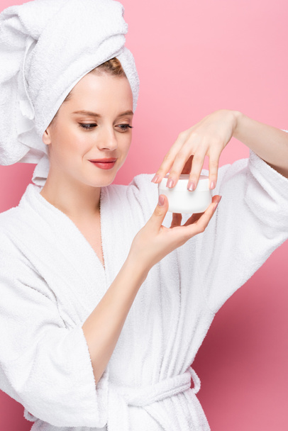 young woman in bathrobe with towel on head opening cosmetic cream isolated on pink - Foto, Bild
