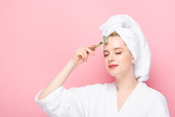 young woman in bathrobe with towel on head using jade roller isolated on pink - Photo, Image