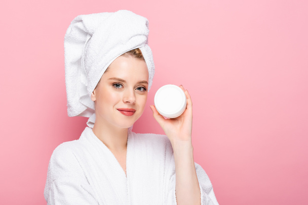 young woman in bathrobe with towel on head holding cosmetic cream isolated on pink - Zdjęcie, obraz