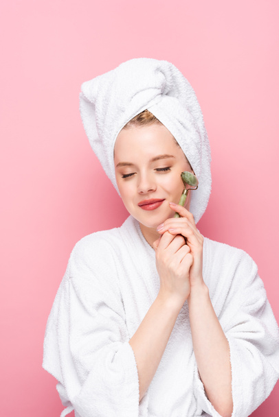 young woman in bathrobe with towel on head using jade roller with closed eyes isolated on pink - Foto, Imagem