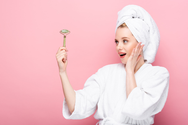 shocked young woman in bathrobe with towel on head holding jade roller isolated on pink - Foto, Imagem