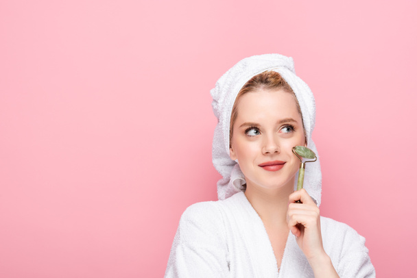dreamy young woman in bathrobe with towel on head using jade roller isolated on pink - Photo, Image