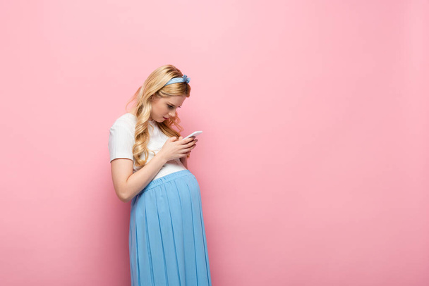 блондинка молода вагітна жінка використовує смартфон на рожевому фоні
 - Фото, зображення