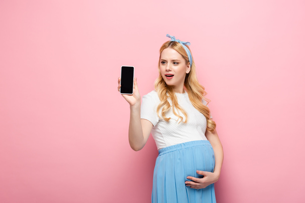 σοκαρισμένη ξανθιά νεαρή έγκυος γυναίκα δείχνει smartphone σε ροζ φόντο - Φωτογραφία, εικόνα