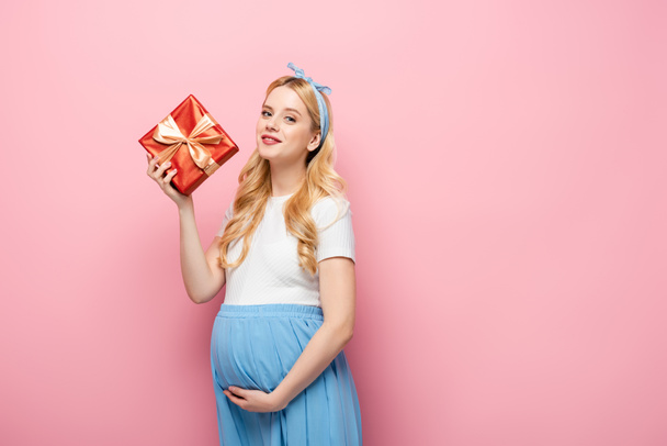 rubia joven embarazada con caja de regalo sobre fondo rosa - Foto, imagen