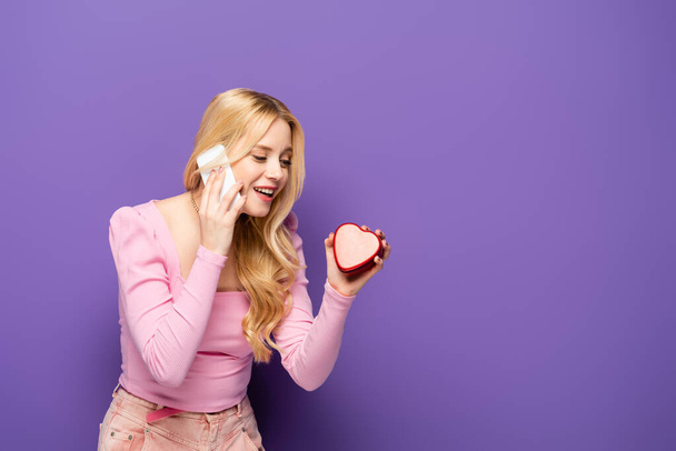 happy blonde young woman with red heart shaped box talking on smartphone on purple background - Foto, Imagen
