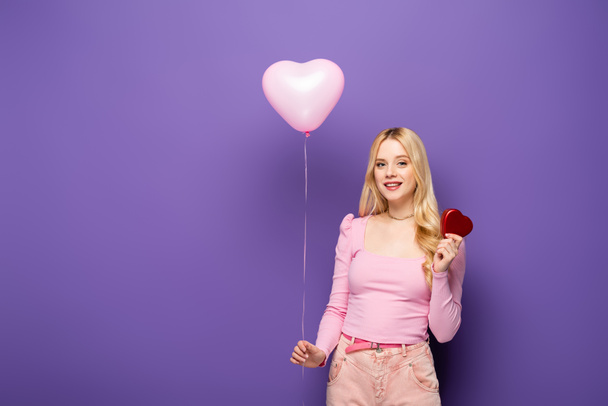 smiling blonde young woman holding heart shaped balloon and gift on purple background - Fotografie, Obrázek