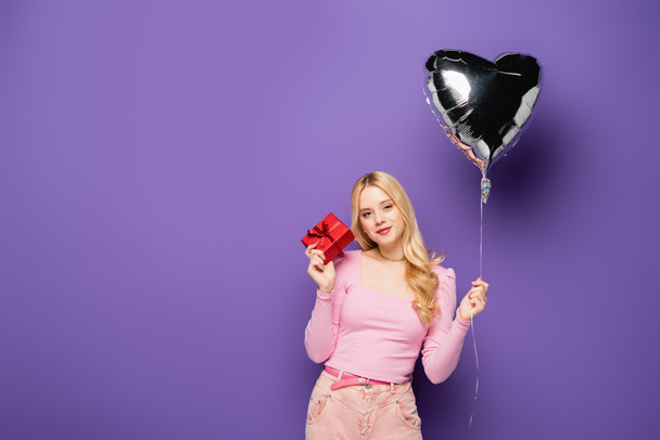blonde young woman holding heart shaped balloon and gift on purple background - Foto, Imagem