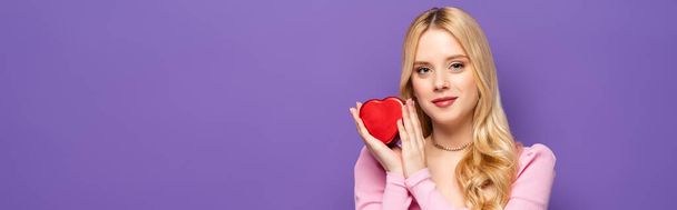 blonde young woman holding red heart shaped box isolated on purple, banner - Foto, Imagen
