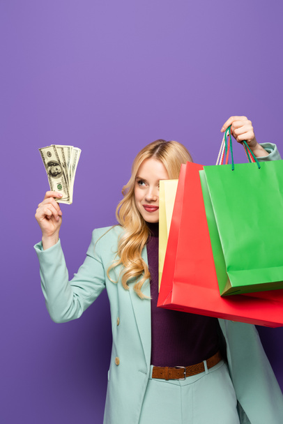 blonde jonge vrouw in trendy turquoise blazer met boodschappentassen en geld op paarse achtergrond - Foto, afbeelding