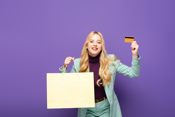 glückliche blonde junge Frau im modischen türkisfarbenen Blazer mit Kreditkarte und Einkaufstasche auf lila Hintergrund - Foto, Bild