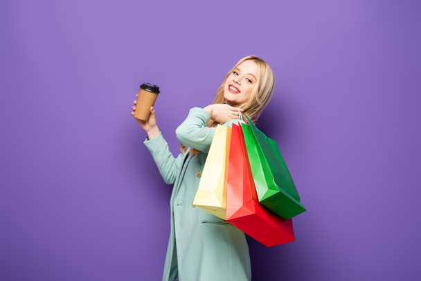 happy blonde young woman in fashionable turquoise blazer with shopping bags and paper cup on purple background - Photo, Image
