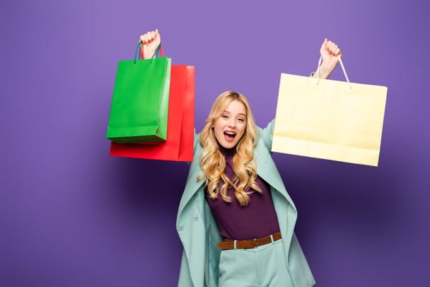 happy blonde young woman in fashionable turquoise blazer with shopping bags on purple background - Photo, Image