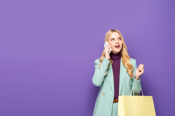 mujer joven rubia en blazer turquesa de moda hablando en el teléfono inteligente y sosteniendo el bolso de compras sobre fondo púrpura - Foto, Imagen