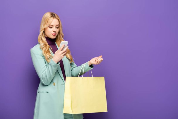 jovem loira em blazer turquesa elegante com smartphone e saco de compras no fundo roxo - Foto, Imagem