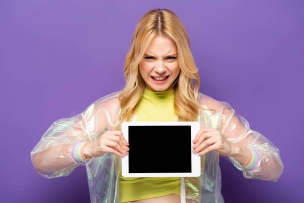 verwirrte blonde junge Frau in buntem Outfit präsentiert digitales Tablet auf lila Hintergrund - Foto, Bild