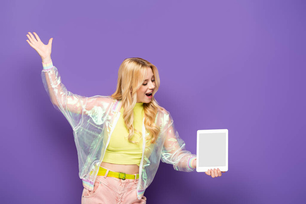 happy blonde young woman in colorful outfit presenting digital tablet on purple background - Fotografie, Obrázek