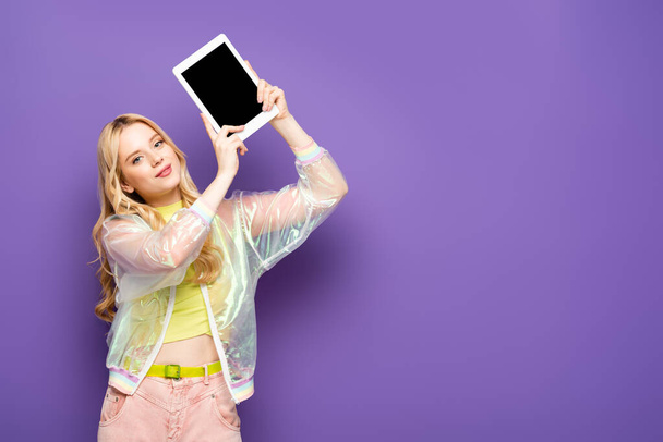 blonde young woman in colorful outfit presenting digital tablet on purple background - Photo, Image
