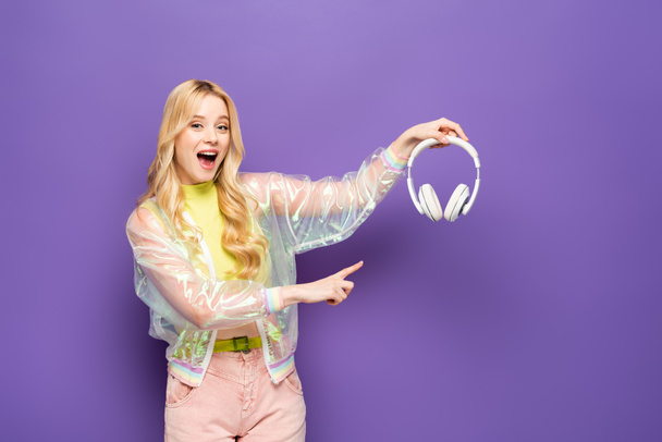 opgewonden blonde jonge vrouw in kleurrijke outfit wijzend op koptelefoon op paarse achtergrond - Foto, afbeelding