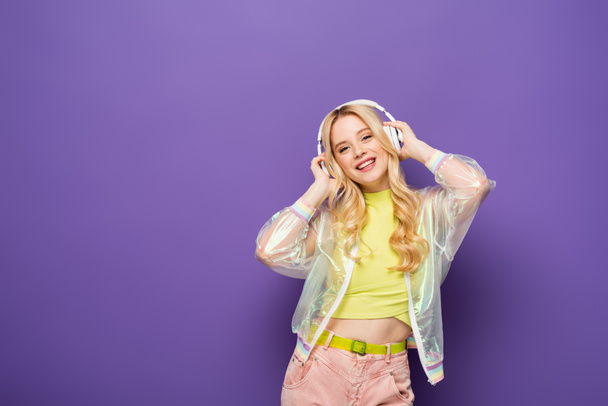 happy blonde young woman in colorful outfit and headphones on purple background - Foto, afbeelding