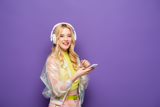 happy blonde young woman in colorful outfit and headphones holding smartphone on purple background - Photo, Image