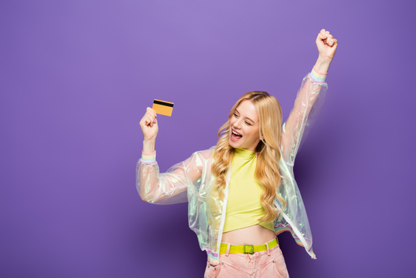 feliz rubia joven en traje colorido mostrando tarjeta de crédito sobre fondo púrpura - Foto, Imagen
