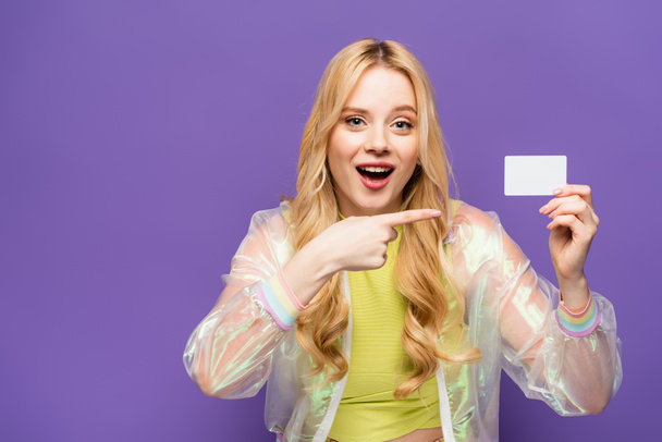 shocked blonde young woman in colorful outfit pointing at blank card on purple background - Photo, Image