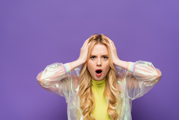 shocked blonde young woman in colorful outfit holding head on purple background - Foto, Imagen