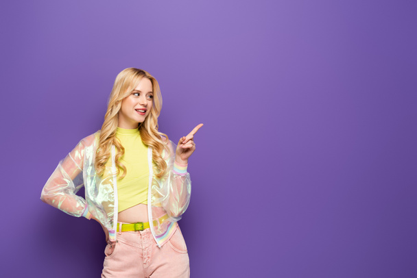 smiling blonde young woman in colorful outfit pointing aside on purple background - Photo, Image