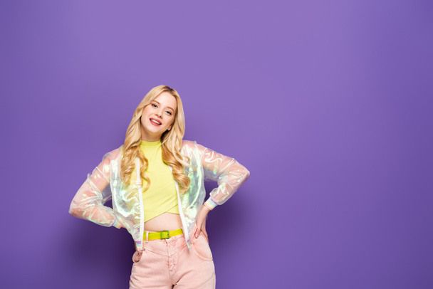 smiling blonde young woman in colorful outfit posing with hands on hips on purple background - Photo, image