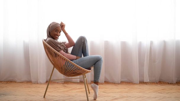 Тисячолітня чорна жінка в хіджабі сидить у зручному кріслі, дивлячись на камеру і посміхаючись вдома, порожній простір
 - Фото, зображення
