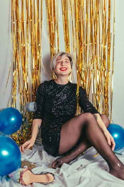 Een jonge blonde vrouw die oudejaarsavond viert. Glitter sprankelende confetti. Cocktailparty avond. Veel plezier. Gouden muur. rode lippen. Luxe jurk. Wit zwart. Blauwe ballonnen. Lachende persoon - Foto, afbeelding