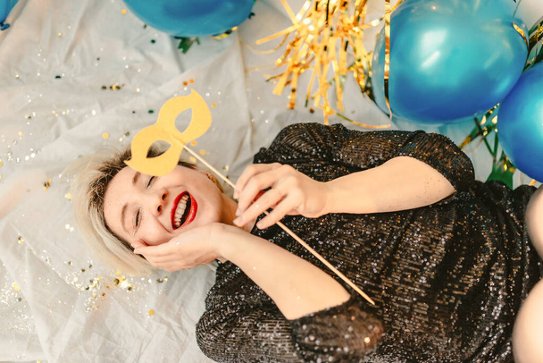 Young millennial blonde woman celebrating New Years Eve. Glitter sparkling confetti. Cocktail party night. Having fun. Golden wall. red lips. Luxury dress. White black. Blue balloons. Smiling person - Photo, image