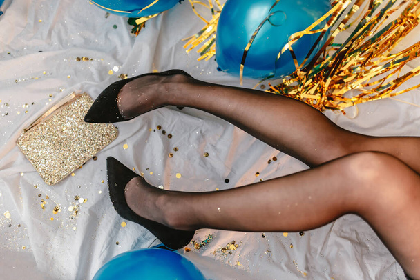 Woman celebrating New Years Eve. Holding black high heels. After party. Glitter sparkling confetti. Cocktail party night. Having fun. Golden wall. red lips. Luxury dress. White black. Blue balloons. - Foto, Imagem