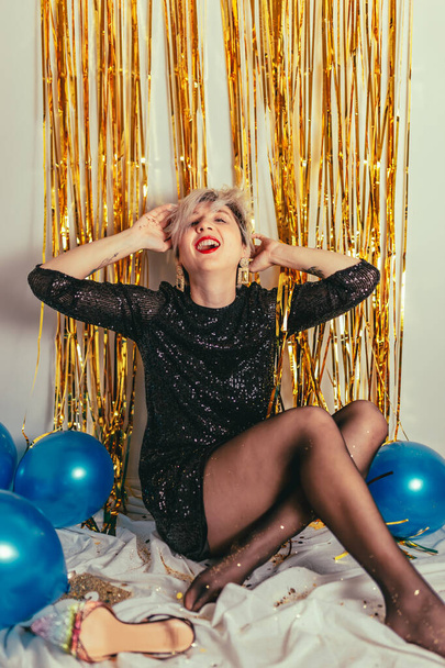 Молода тисячолітня блондинка святкує Новий рік. Блискуча ігристий конфетті. Вечірка з коктейлем вночі. Розважатися. Золота стіна. червоні губи. Розкішне плаття. Білий чорний. Блакитні кульки. Усміхнена людина
 - Фото, зображення