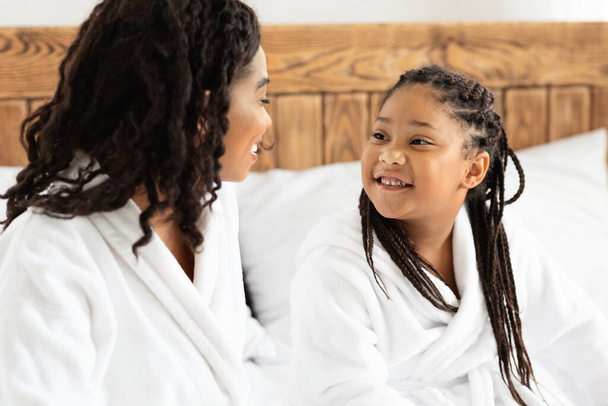 Linda niña negra y su mamá charlando en la cama después del baño - Foto, Imagen